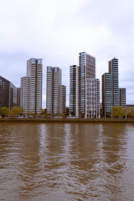 A row of modern high-rise apartment buildings is visible along the riverbank against a partly cloudy sky near Surrey Quays. The buildings feature balconies and a variety of geometric windows, with som